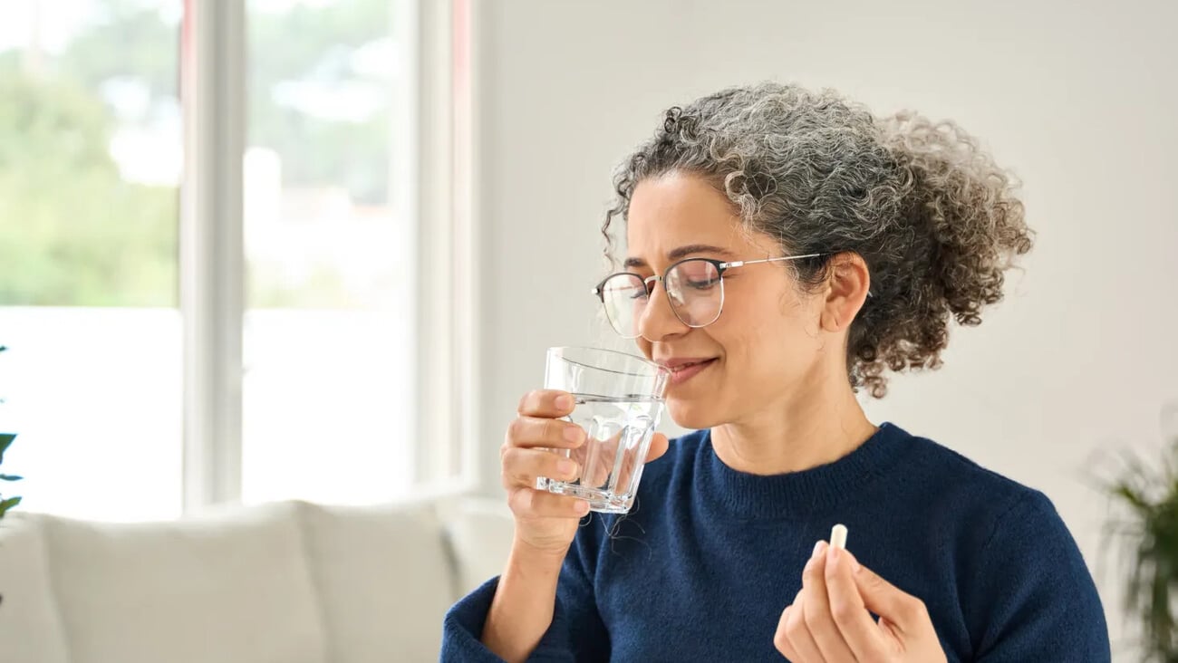 Happy healthy woman of middle age talking pill holding glass of water standing at home. Mature 50 years old lady taking vitamin health care supplement morning daily nutrition treatment concept.; Shutterstock ID 2516642939