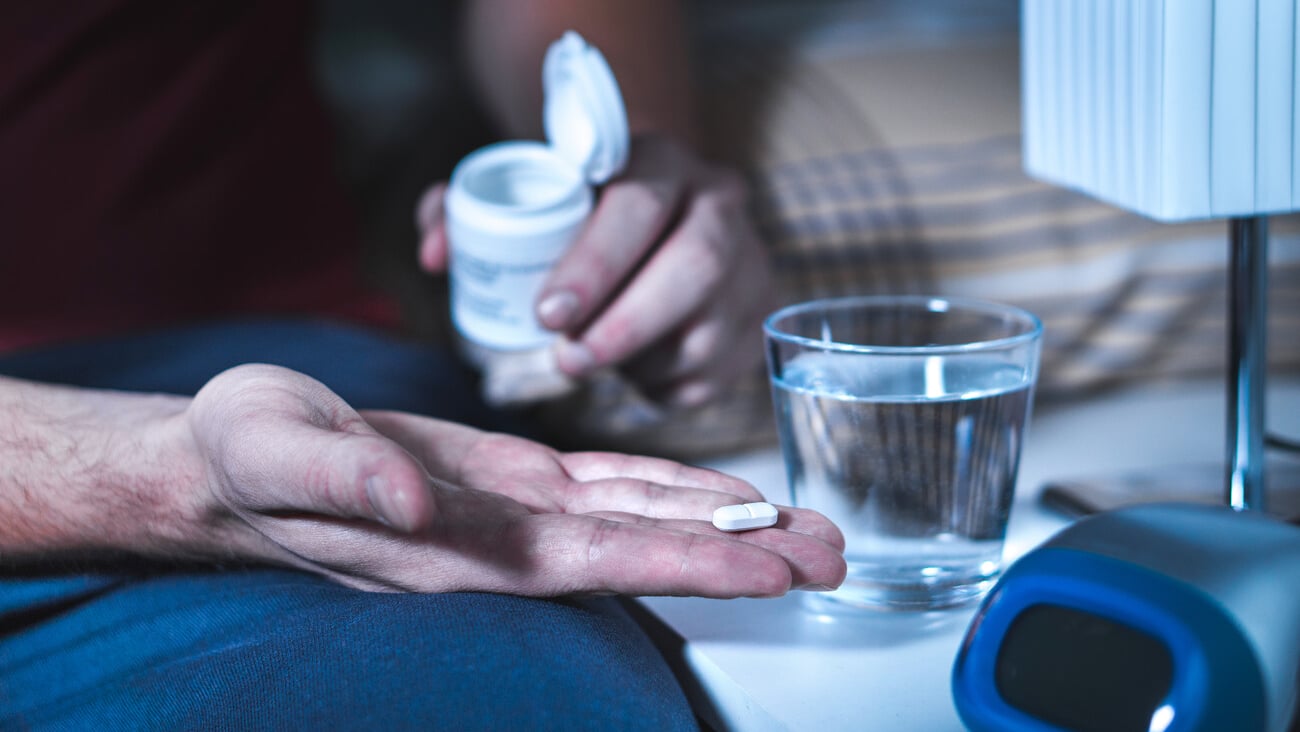 Man sitting on bed about to take sleeping pill or night medicine. Suffering from insomnia. Holding tablet and bottle in hand. Glass of water on nightstand in bedroom.; Shutterstock ID 756787537