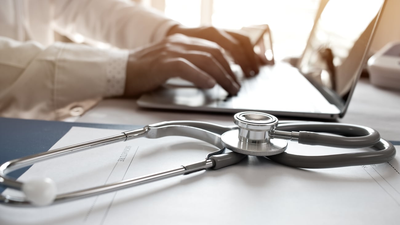 A physician's hands typing on a laptop with a stethoscope in the foreground