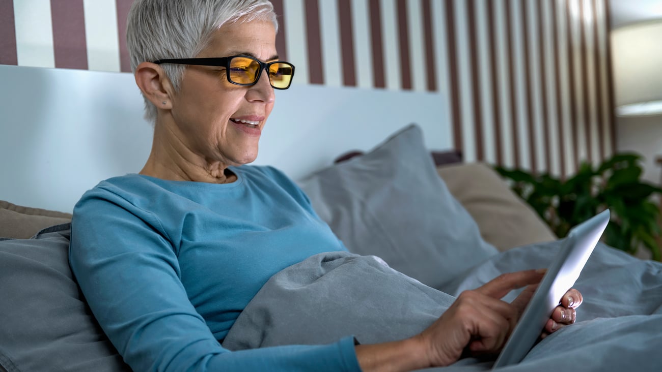 Woman with short grey hair wearing glasses and reading tablet in bed