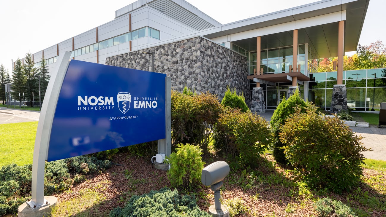 A blue sign saying NOSM is outside a modern education building about four stories high.
