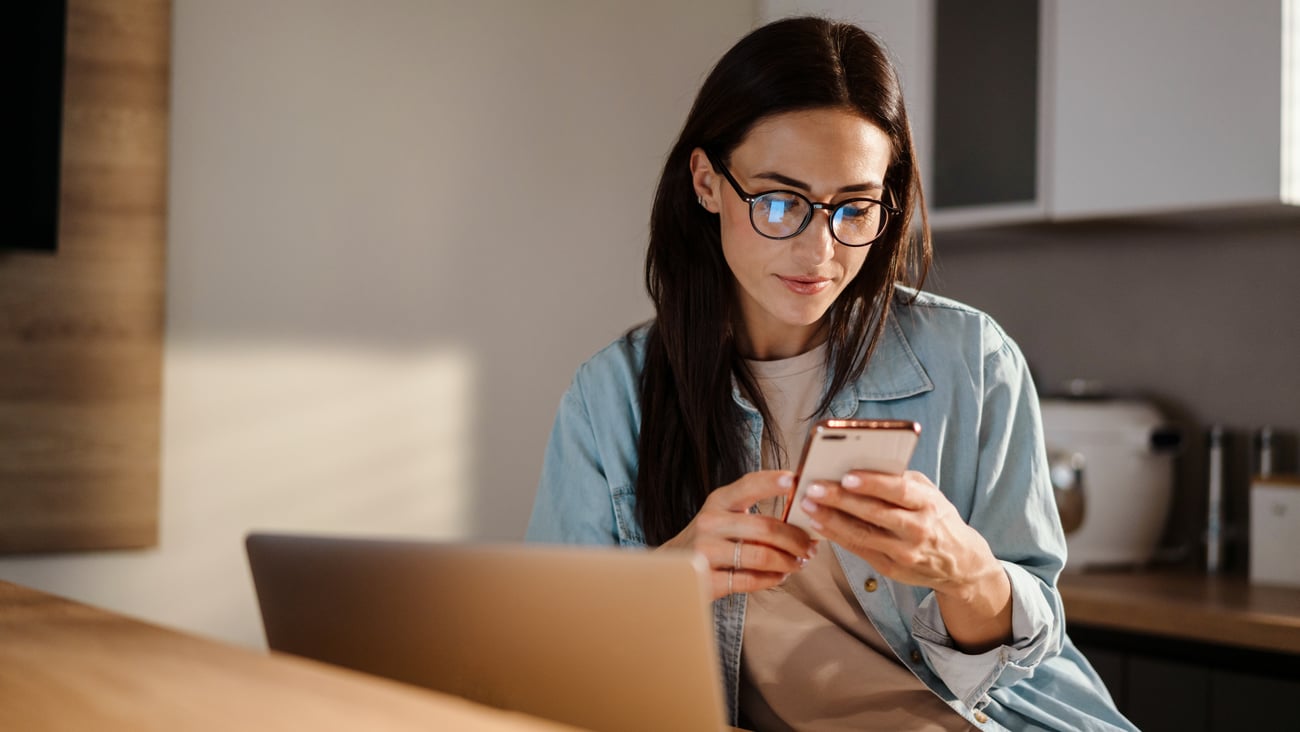 a woman sitting at her laptop, looking at cellphone