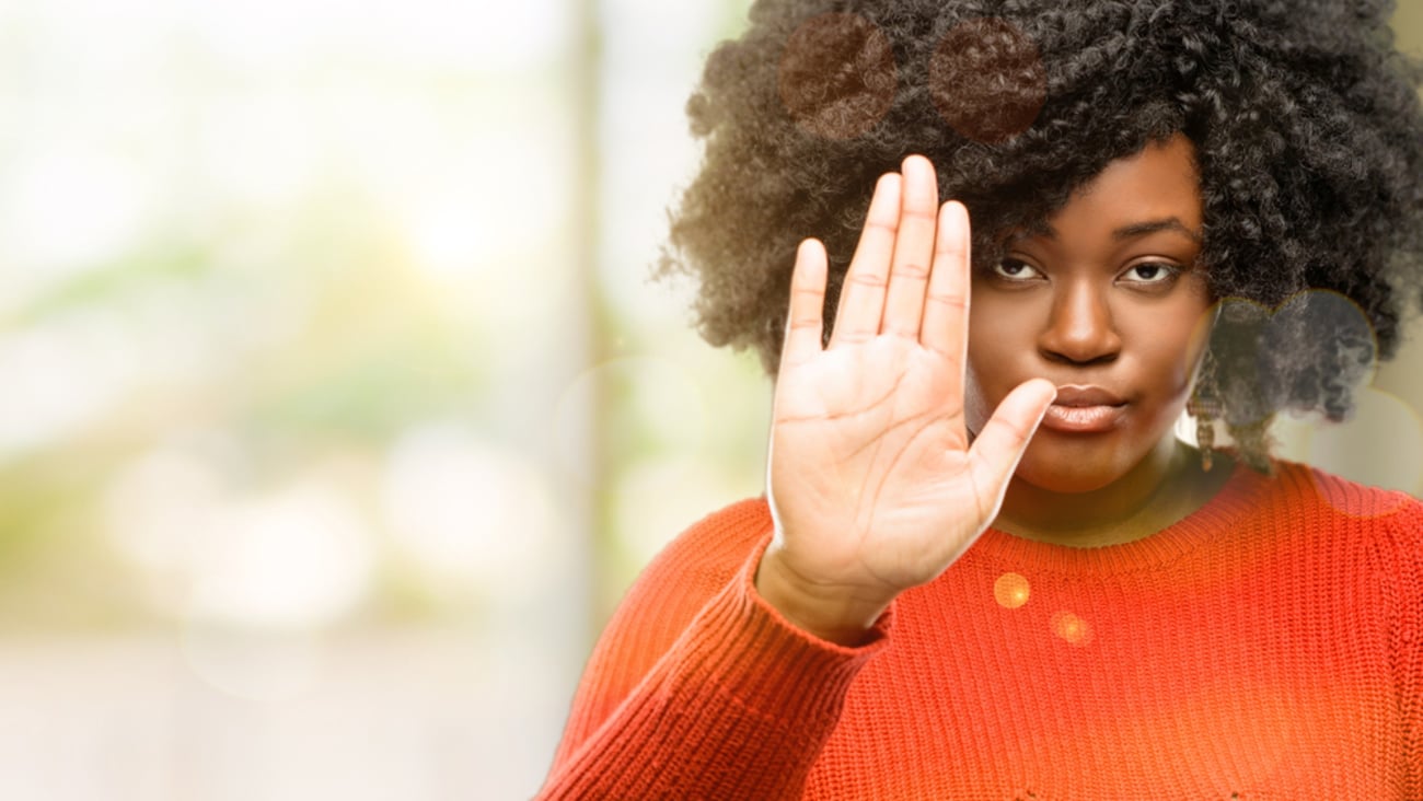 Black woman in orange sweater holding up her hand to say no