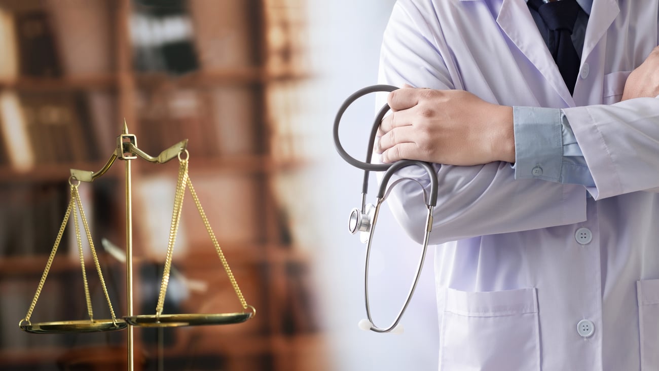 Torso of doctor in white coat holding stethoscope beside gold justice scales