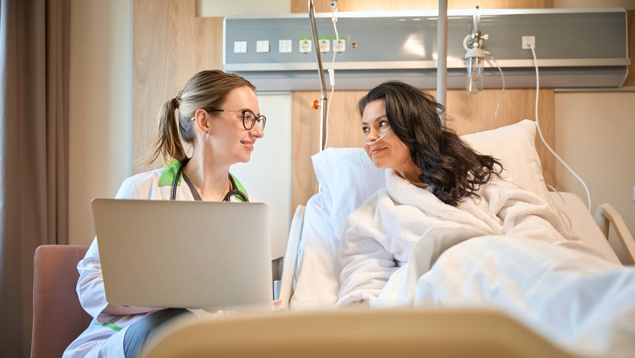 A doctor sits at a chair speaking to a patient at a hospital bed.