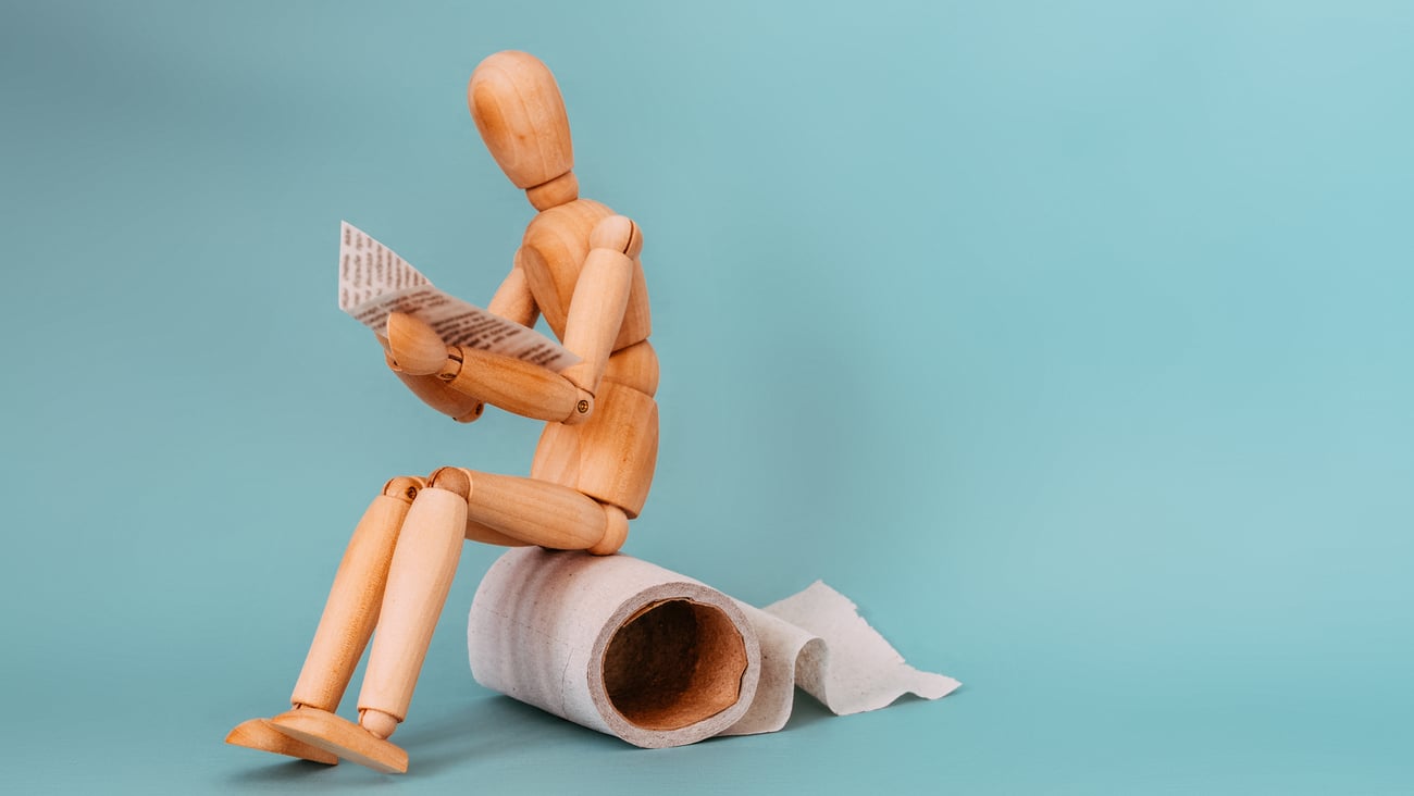 Wooden figure sitting on roll of toilet paper, reading newspaper