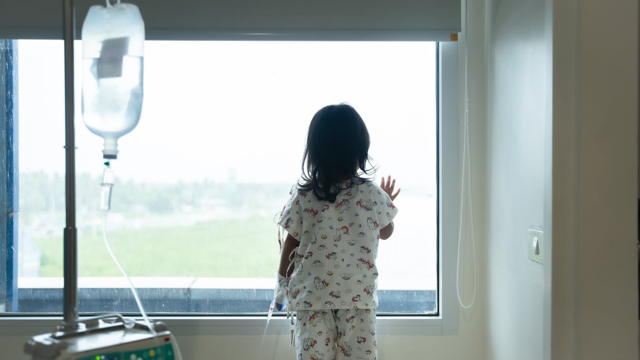 Child wearing IV tube is standing and looking out the window in hospital