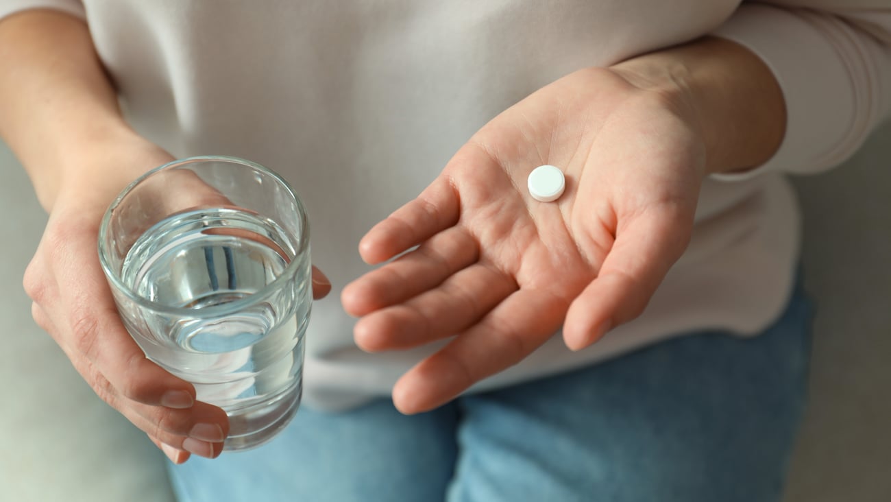 white woman, sitting, holding round white pill in one hand and glass of water in the other, image just showing torso, 