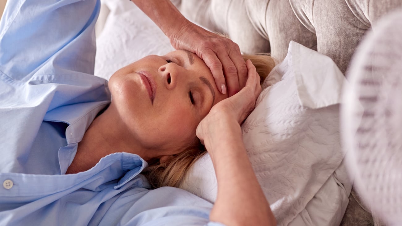 Woman in bed holding forehead with eyes closed and fan on her, suggesting hot flush