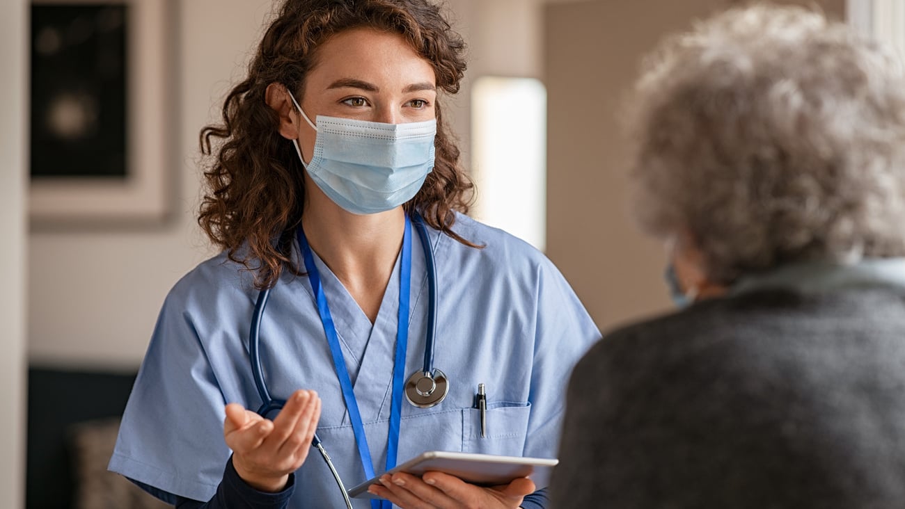 Doctor wearing safety protective mask supporting and cheering up senior patient during home visit during covid-19 pandemic. Nurse and old woman wearing facemasks during coronavirus and flu outbreak. ; Shutterstock ID 1852862014