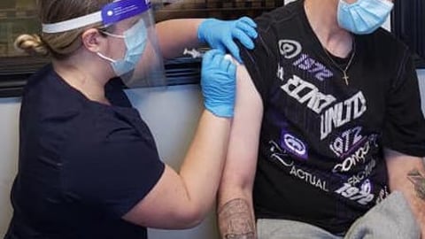 Pharmacist Janice Audeau wears a mask and face shield and black uniform as she gives a vaccine to a masked male patient in his 30s.