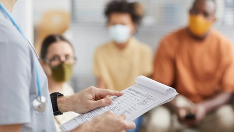 A male physician holds a clipboard in a waiting room with patients wearing masks.