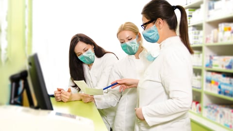 Three female pharmacists wearing lab coats and masks stand behind a pharmacy counter looking at documents.