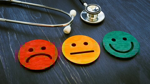 A stethoscope sits on a table with three wooden round emojis; one sad, one neutral and one smiling