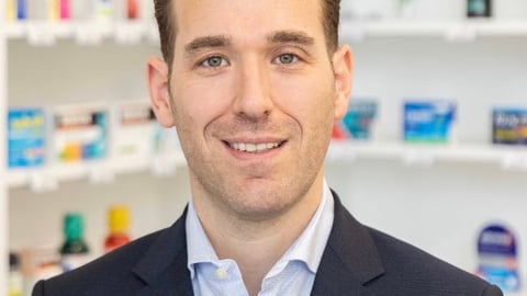 Head shot of Adam Silvertown in a blue suit with pharmacy shelves behind him