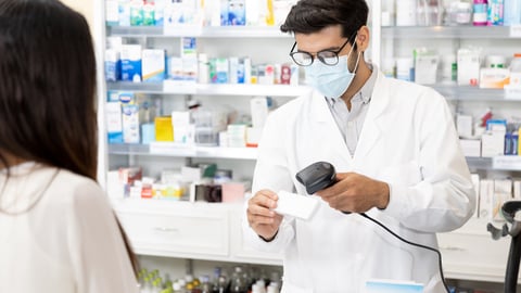 A male pharmacist wearing a mask uses a hand-held scanner to process the cost of medication at a pharmacy counter while a female patient waits.