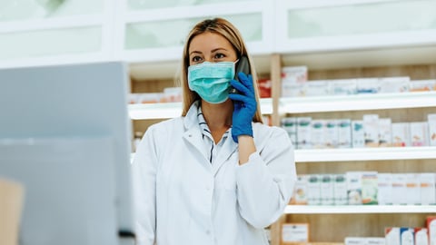 A female pharmacist wearing a mask stands behind a pharmacy counter speaking on the phone.