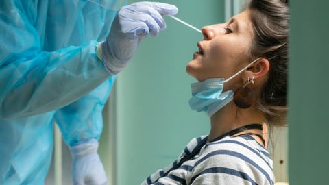 A woman sits while being tested for COVID with a nasal swab, held by a  gowned person with gloves on.