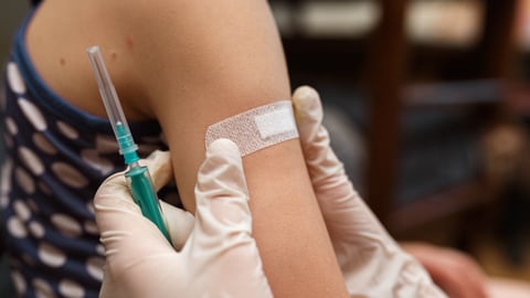 Photo of a gloved healthcare worker holding a needle and putting a bandage on a young child's arm. 