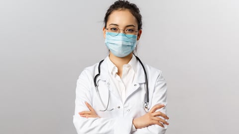 A young female physician wears glasses, a mask a lab coat and has a stethoscope around her neck.