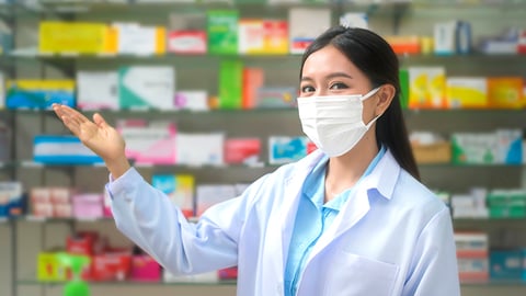 young female pharmacist wearing a mask, arm extended toward shelves of products