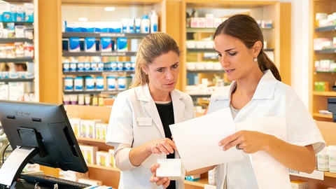 an experienced female pharmacists instructs a younger female pharmacy technician on how to check paperwork