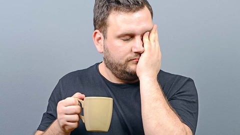 sleepy 30-something man holding a cup of coffee and rubbing his face