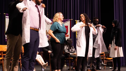 three medical students put on lab coats while smiling mentors look on