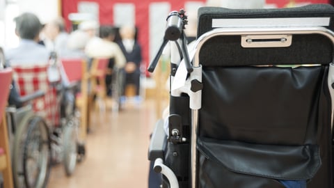 The back of a wheelchair in the foreground with blurry seniors resting in the background