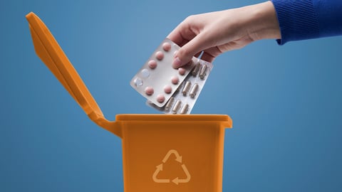 A hand throwing medications into a tiny orange trash can 