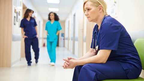 Female nurse looking tired, pensive, stressed, with two other female nurses chatting in the background