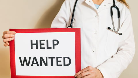 an anonymous female physician holds a sign reading “help wanted”