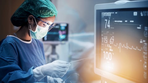 Physician in a hospital room with hairnet and mask on standing next to a heart monitor