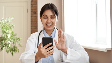 Young woman South Asian doctor sitting at a desk talking to a patient over video chat on her phone