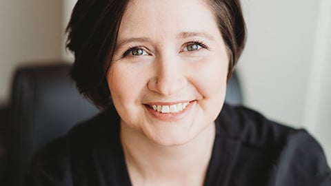 a Caucasian woman with short dark hair smiles at the camera. She is wearing a dark sweater