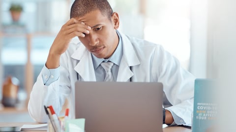 Young male Black doctor looking at his computer with his head in one hand in frustration