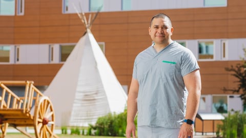 Tall man wearing scrubs outside a hospital in front of a traditional Indigenous structure