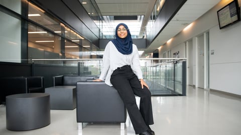 A brown woman wearing a hijab, white dress shirt and black pants leans against a chair