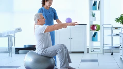 white-haired man sitting on exercise ball being assisted by physiotherapist