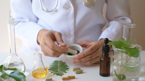 scientist or doctor with mortar and pestal mixing herbs, with supplement pills on table and a vial and green plant