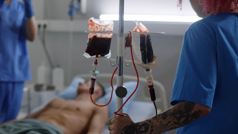 young white male patient in hospital bed getting blood transfusion, blood bags hanging, healthcare professionals standing by 