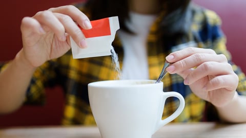 Close up of a young person pouring a packet of sugar substitute into a cup of coffee