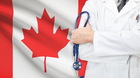 Torso of male doctor in white coat standing in front and to the side of Canadian flag