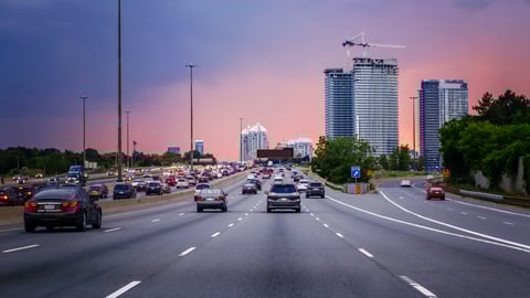 Image of cars on 3-lane road with high rise buildings in the background and pinky hazy sky.