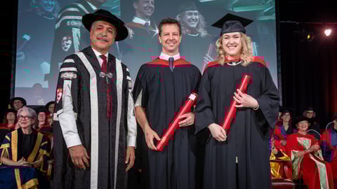 The president and vice-chancellor of McGill wearing academic robes and cap stands next to two physicians in academic robes and mortar boards, each clutching a red tube