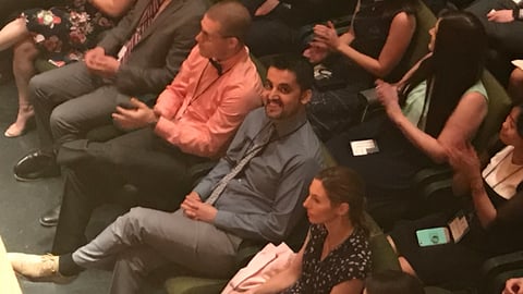 A man sitting in a large audience, smiling up at someone in the balcony