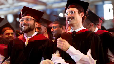 A man with a beard and a man with a mustache sit smiling beside each other wearing academic robes at a convocation ceremony.