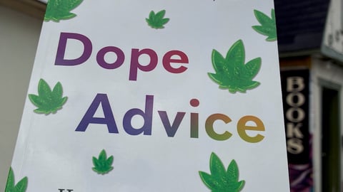 a copy of the paperback book Dope Advice is held up against a cloudy sky with a cute book store in the background