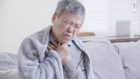 An elderly asian man in grey sweater clutches his sore throat