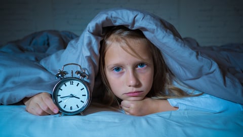 Young girl unable to sleep, under her covers peeping out with an alarm clock